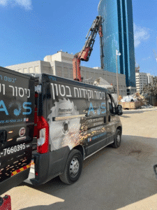 רכבי שירות ממותגים של חברת ניסור וקידוח בטון חונים באתר עבודה על רקע מנוף ומגדל משרדים.